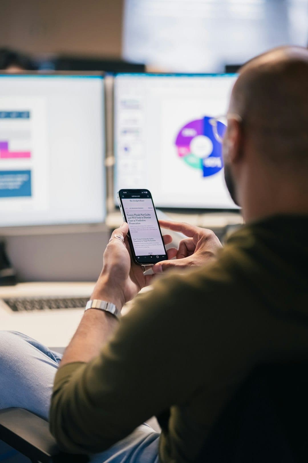 A man reviewing data on a smartphone in front of computer monitors